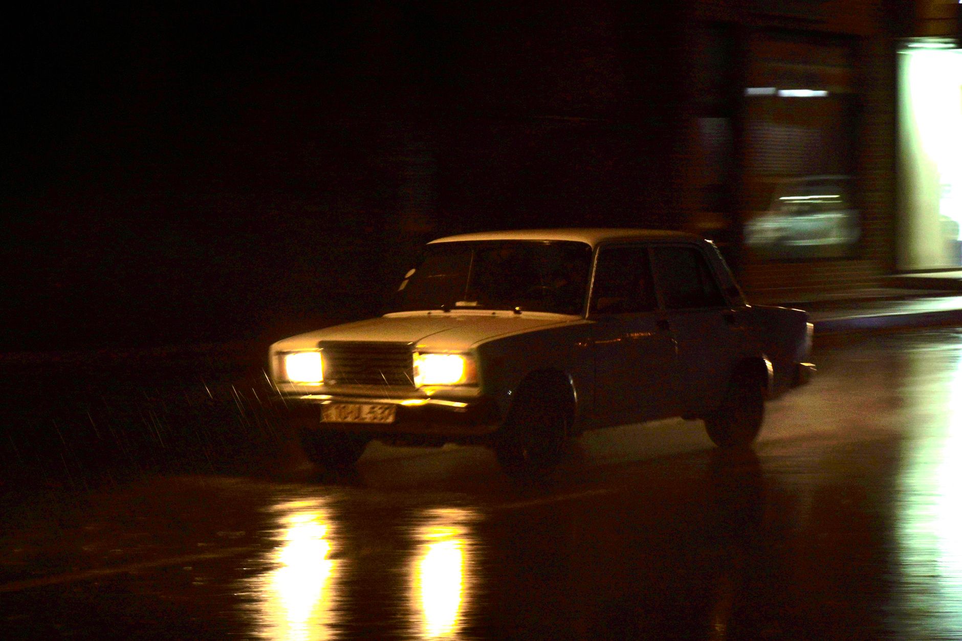 A car on a wet street at night with headlights and reflections spread across the road surface